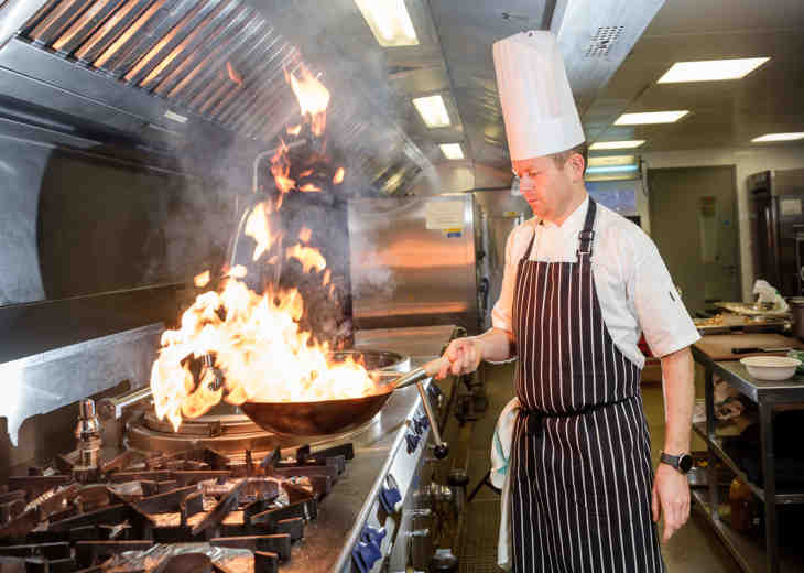 A chef standing a kitchen whilst holding a flaming frying pan.