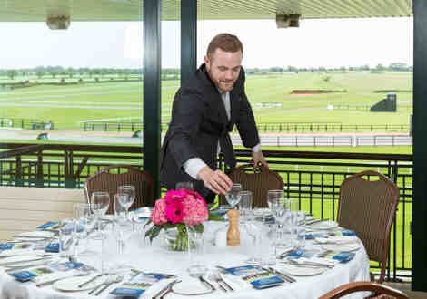 A man arranging glasses on a dining table in a racecourse venue.